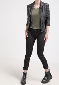 Giacca da biker in pelle nera con dettagli trapuntati sulle spalle, camicia aderente verde oliva e jeans skinny neri arrotolati alle caviglie, abbinati a stivali neri voluminosi.