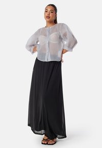 Blusa corta semi-trasparente di colore azzurro chiaro con maniche a sbuffo e quattro grandi bottoni, abbinata a una lunga gonna nera fluida.