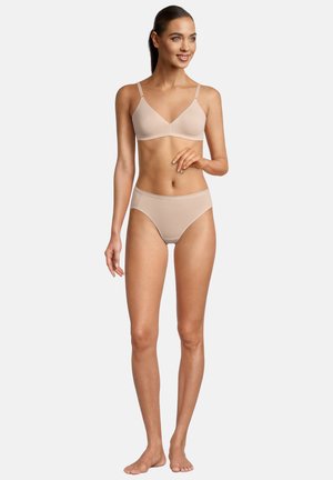 Schlanke Frau steht barfuß, trägt einen beigen kabellosen BH und hoch taillierte Höschen, lächelt mit der rechten Hand in Hüfthöhe und der linken Hand entspannt.