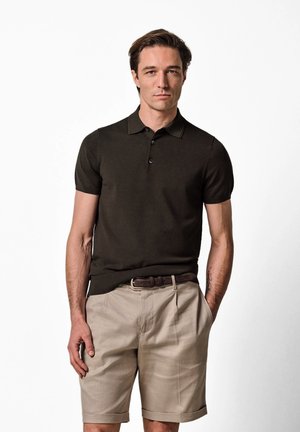 Mężczyzna w ciemnobrązowej, krótkiej koszulce polo i beżowych szortach do kolan, stoi z jedną ręką w kieszeni na białym tle.