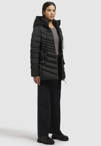 Sort puffet jakke med hætte, med quiltet tekstur, justerbare snore og en sidelomme. Sættes sammen med sorte bukser og støvler.