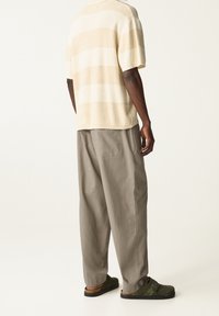 Beige en crème gestreepte gebreide shirt, losse grijze broek en donkergroene instappers. Casual, relaxed fit met zichtbare textuur en contrasterende kleuren.