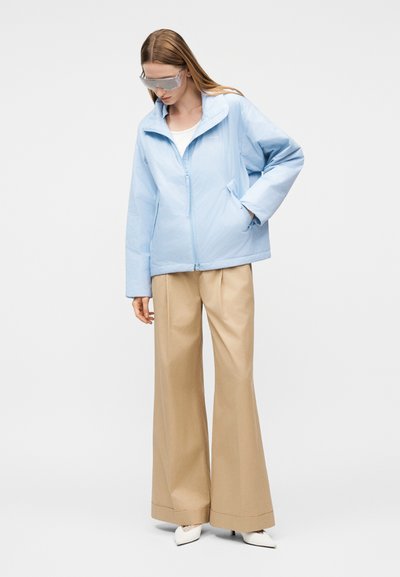 Γυναίκα που φοράει ανοιχτό μπλε μπουφάν με φερμουάρ, μπεζ παντελόνι φαρδύ, λευκά μυτερά παπούτσια και διάφανα προστατευτικά γυαλιά, στέκεται με το ένα χέρι στην τσέπη.