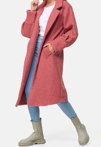 Manteau long texturé rose avec devant ouvert, poches latérales et col cranté, accompagné d'un jean bleu clair et de bottines gris clair.