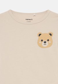 Beige cotton t-shirt featuring a bear face graphic on the upper left side. Round neck and soft texture. No visible patterns or accents.