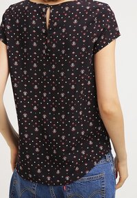 Černá blůza s krátkými rukávy, s květinovým vzorem v červené a bílé barvě. Detail na zádech s klíčovou dírou a knoflíkovým zapínáním. Mírně zakřivený lem.