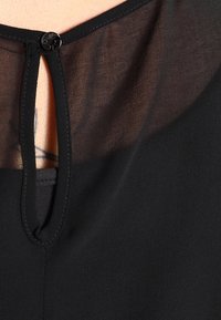 Černá blůza s průhlednou, strukturovanou částí u výstřihu, která je zajištěna kulatým černým knoflíkem a malým otvorem ve tvaru klíče.