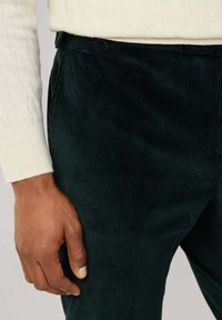 Tmavozelené kordové nohavice nosené s bielym svetríkom s pleteným vzorom, zobrazenie detailu ruky osoby odpočívajúcej popri tele.