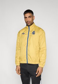 Yellow lightweight jacket with blue zip and accents, featuring high collar and embroidered logo on the chest. Smooth texture, regular fit.