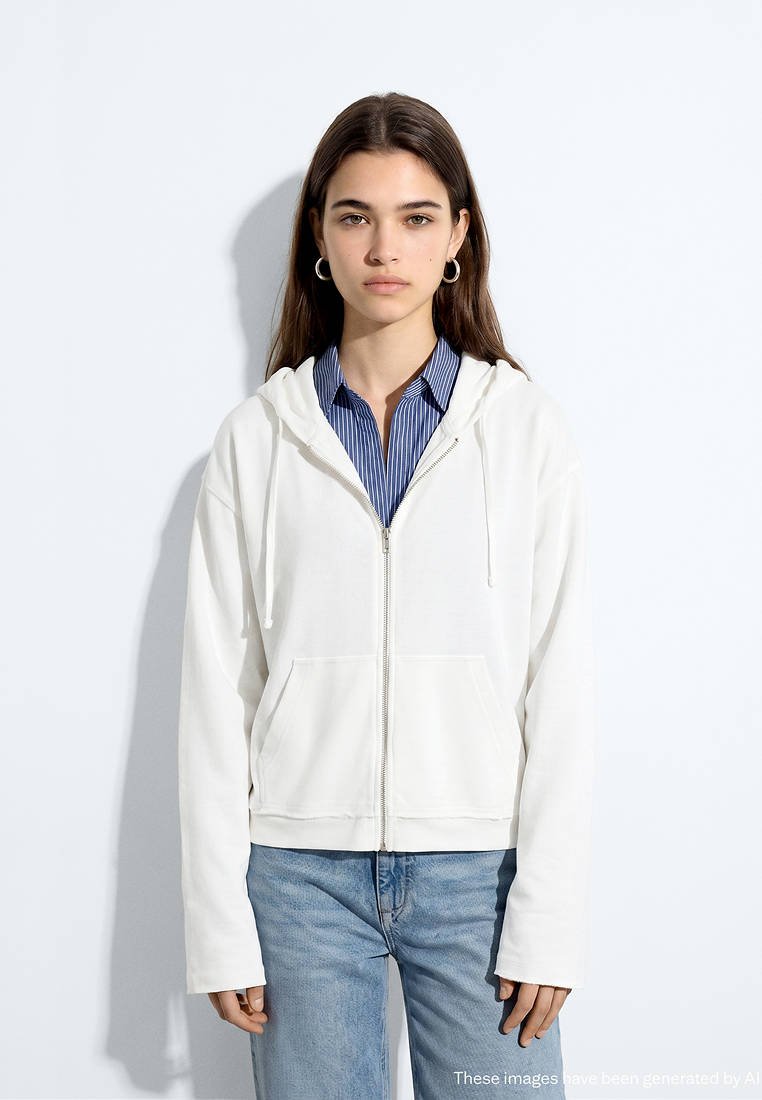Jeune femme aux longs cheveux bruns porte des boucles d'oreilles créoles, un sweat à capuche blanc zippé sur une chemise rayée bleue et un jean bleu clair, debout contre un mur blanc.