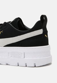 Černé semišové tenisky s bílou gumovou podešví, které mají texturovanou střední podešev, kontrastní šedý pruh a zlaté logo Puma na boku.