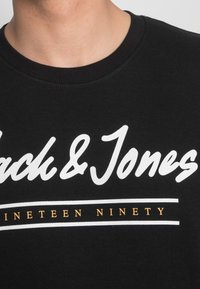 Sudadera negra con texto en cursiva blanco "Jack & Jones" y la frase "Nineteen Ninety" en naranja debajo, con un cuello acanalado.