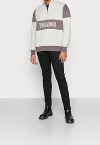 Sudadera bloqueada en gris y blanco con cuello con cremallera, que presenta el texto "PEGADOR"; combinada con jeans negros ajustados y botas negras.