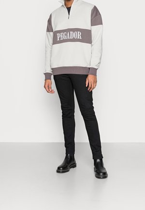 Grå og hvid farveblokeret pullover med lynlås krave, med "PEGADOR" tekst; parret med sorte slim-fit jeans og sorte støvler.