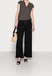 Svarta vida byxor i kombination med en mönstrad blus med kort ärm. Tillbehören inkluderar en liten vävd handväska och svarta slidesandaler.