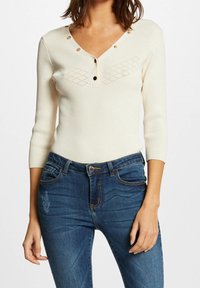 Langärmeliges, elfenbeinfarbenes Ripp-Top mit V-Ausschnitt, dekorativen goldenen Knöpfen und dezenten Diamantmustern auf der Brust, kombiniert mit blauen Jeans.