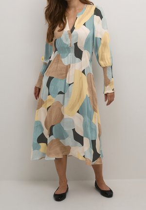 Kvinde iført en langærmet midi-kjole med abstrakt multicolor mønster i beige, blå, gul, sort og hvid, kombineret med sorte flade sko.