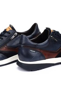 Tummansinisiä ja ruskeita nahkasneakers-kengät, joissa on valkoinen kumipohja, teksturoidut yksityiskohdat ja sivu vetoketju. Sääntöä brändäys kantapäässä ja kielekkeessä.