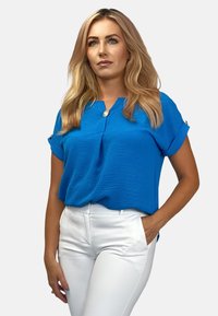 Niebieska bluzka krótki rękaw o fakturowanej powierzchni, z kołnierzykiem typu notch i ozdobnym detalem w postaci guzika, połączona z białymi dopasowanymi spodniami.