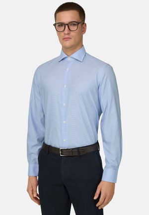 Mężczyzna w czarnych okularach, jasnobłękitnej koszuli zapinanej na guziki, włożonej do czarnych spodni z brązowym paskiem, stojący na białym tle.