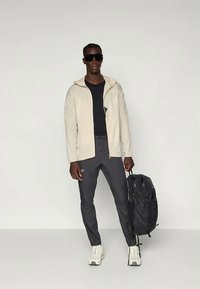 Svetlo bež jakna na zadrgo, črna majica, temno sivi hlače, beli superge in črn nahrbtnik. Sončna očala dopolnjujejo videz.