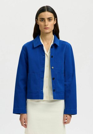 Kvinne med langt mørkt hår iført en knallblå knappet jakke over en hvit bluse og skjørt, stående foran en ensfarget lys bakgrunn.