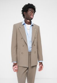 Double-breasted beige blazer with silver buttons, paired with a light blue shirt and white undershirt, worn with matching trousers.
