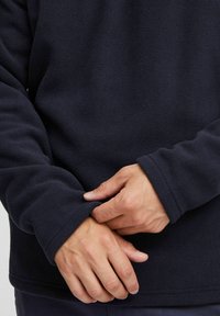 Marinblå fleece-tröja med en mjuk textur, med långa ärmar och en avslappnad passform. Mudden är något rullad vid handlederna, vilket framhäver tyget.