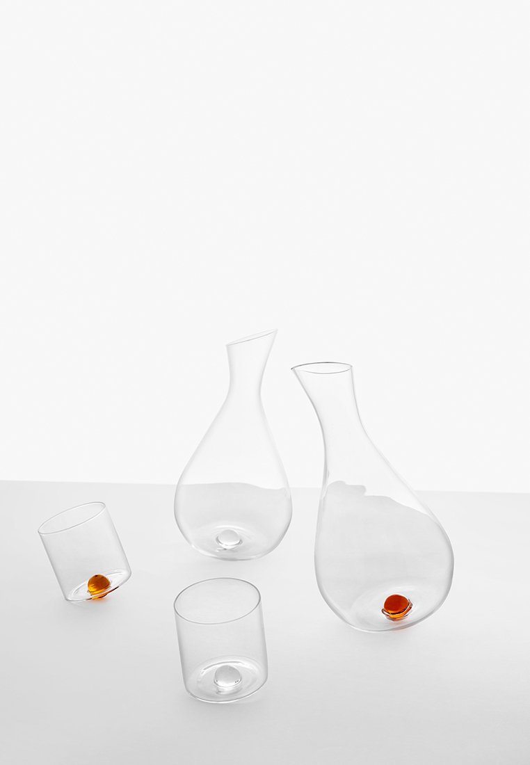 Przezroczysta szklana karafka i dwa szklanki, w kształcie kropli i cylindrycznym, z okrągłymi pomarańczowymi akcentami w każdej podstawie. Gładka tekstura.