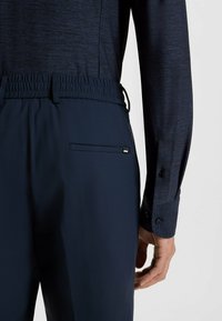 Pantaloni blu navy con vita elastica, tasche laterali e una texture liscia; abbinati a una camicia scura a maniche lunghe con polsini a bottoni.