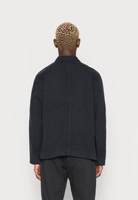 Mörk marinblå jacka i enfärgat tyg, strukturerad form och med en kavajkrage, utrustad med en söm baktill och subtila detaljer vid fållen.