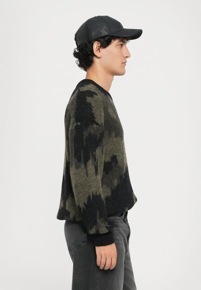 Trui in groen en zwart, versierd met een abstract patroon. Rib gebreide manchetten; gecombineerd met donkergrijze jeans en een zwarte leren pet.