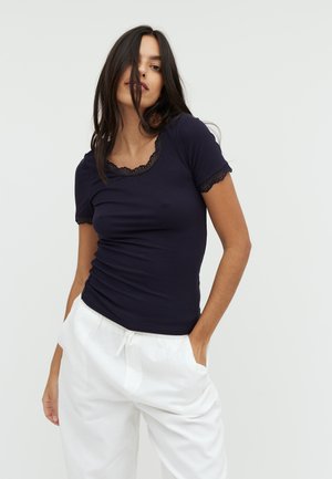 Navy ribbed t-shirt with short sleeves and lace trim at the neckline and sleeve edges, worn with white loose-fit trousers.
