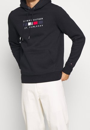 Mężczyzna w czarnej bluzie z kapturem Tommy Hilfiger z logo i białych spodniach, z jedną ręką w kieszeni, na jednolitym tle.