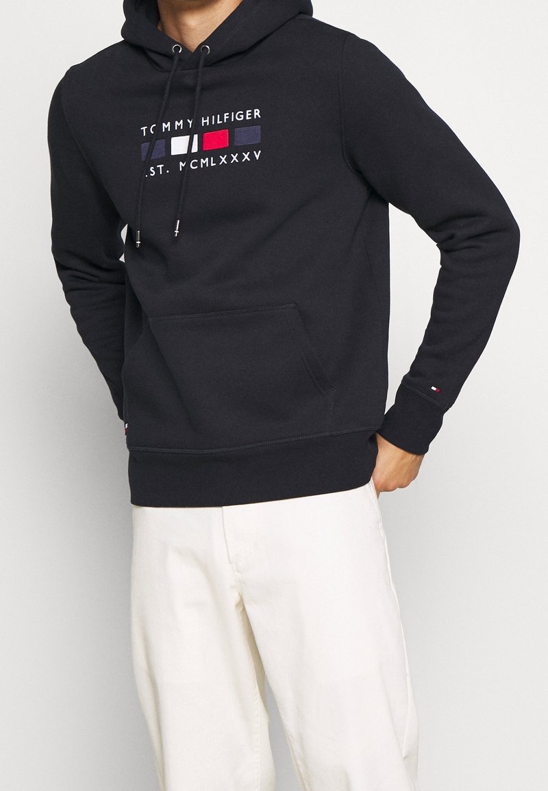 Homme portant un sweat à capuche noir Tommy Hilfiger avec logo et pantalon blanc, une main dans la poche, sur fond uni.
