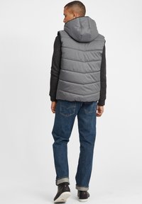 Graue gepolsterte Weste mit Kapuze, mit horizontaler Steppung, getragen über einem schwarzen Pullover, kombiniert mit blauen Jeans und schwarzen Schuhen.