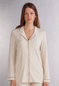 Beige long-sleeve pyjama top met een eventueel geknipt kraag, knopen aan de voorkant en contrasterende bruine piping langs de kraag en manchetten. Zachte stoftextuur.