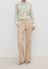 Blusa leggera e trasparente di beige con un motivo floreale in verde e blu tenue; abbinata a pantaloni beige su misura e décolleté neutri a punta.