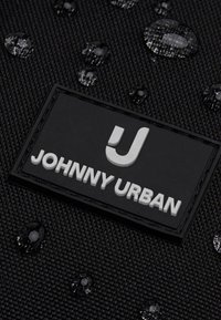 Tela negra texturizada con gotas de agua y una etiqueta rectangular negra que muestra una "J" blanca en relieve y el texto "JOHNNY URBAN".