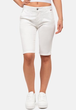 Mujer con pantalones cortos blancos hasta la rodilla y zapatillas blancas, de pie con una mano en la cadera contra un fondo liso.