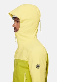 Man draagt een geel tweekleurig capuchonjack met een rits aan de voorkant en het Mammut-logo op de linkerarm, getoond in profiel voor een effen achtergrond.