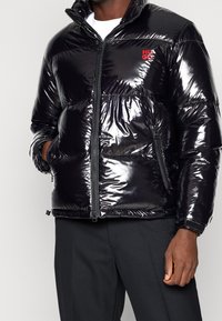 Czarna kurtka puchowa o błyszczącej powierzchni, z zamkiem błyskawicznym z przodu, wysokim kołnierzem oraz czerwonym logo "HUGO" na piersi. Posiada kieszenie boczne.