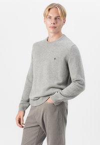 Grå tröja med stickad textur, rund halsringning, ribbade ärmslut och nederkant; har en liten svart logotyp på bröstet. Bärs med beige byxor.