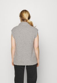 Pull en tricot sans manches gris avec un col montant, présentant un design texturé à côtes et un ourlet arrière plus long, porté avec un pantalon noir.