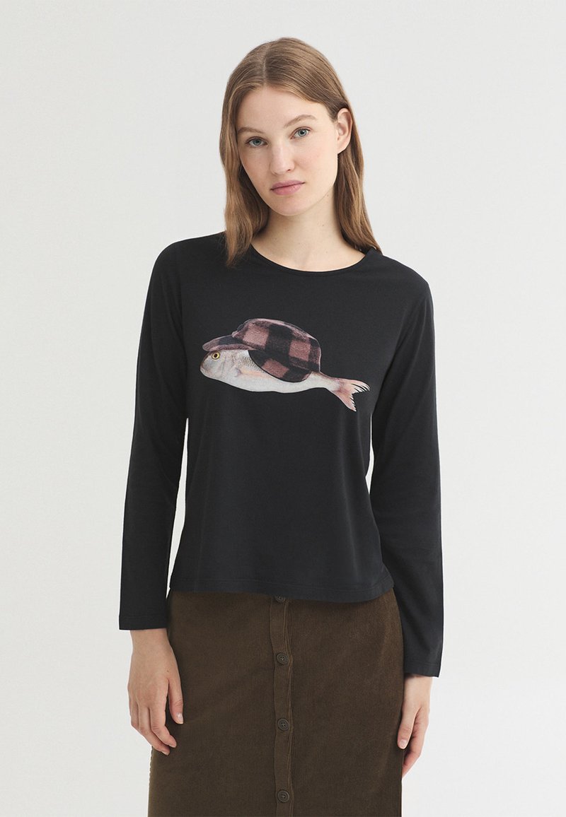 Jeune femme aux cheveux lisses châtain clair porte un haut noir à manches longues avec un motif poisson portant une casquette à carreaux et une jupe marron à boutons.