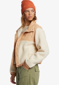 Sherpa bunda s hnedým nylonovým vrchom, krémovým fleece dolným dielom a rebrovanými manžetami. Model má na sebe oranžovú pletenú čiapku. Viditeľné sú aj zelené cargo nohavice.