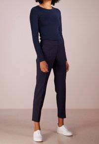 Person, die ein navyblaues langärmeliges Oberteil, navyblaue hochtaillierte Ankle-Hosen und weiße Schnürsneaker trägt, steht auf einem beigen Boden.