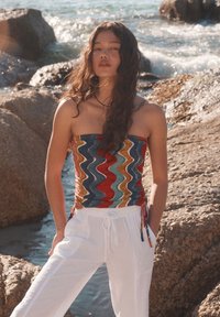 Top senza spalline con motivo a onde colorato abbinato a pantaloni bianchi larghi. Il materiale sembra avere una texture, con una vestibilità comoda. Sullo sfondo ci sono rocce e acqua.
