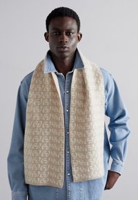 Beige uldskjern med et gentagende logo-mønster, draperet om halsen på en person iført en denimskjorte. Blød tekstur, moderne design.