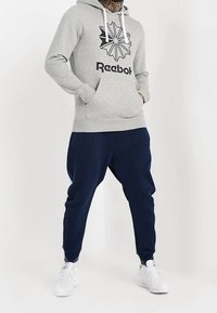 Šedá mikina s kapucí a černým logem Reebok a geometrickým designem, kombinovaná s námořnickými jogger kalhotami a bílými sportovními botami. Hladká látka, casual střih.